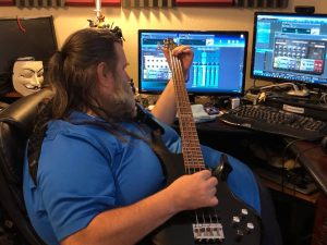 A man is playing an electric bass guitar while looking at music mixing software on his computer monitors